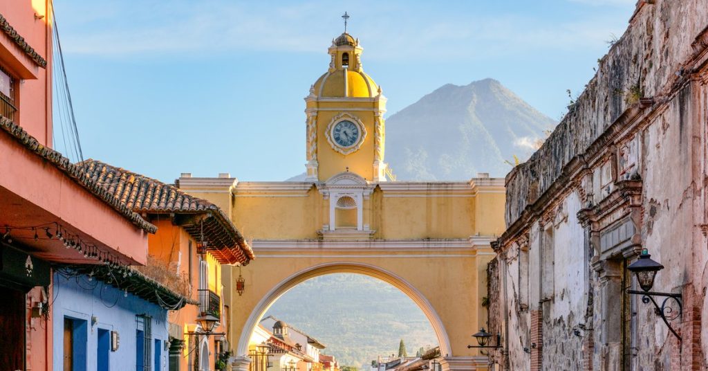 Arco de Santa Catalina sobre la 5.ª avenida Norte de Antigua Guatemala, con su torre del reloj amarilla y el volcán de Agua al fondo